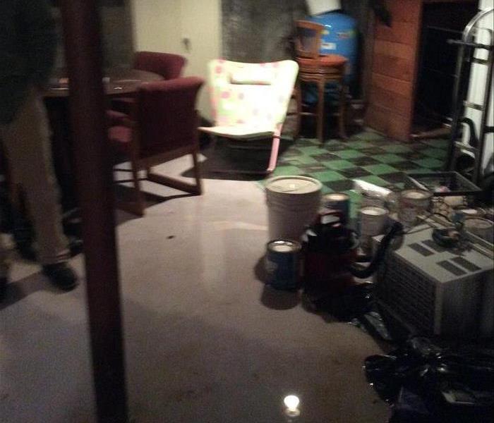 Unfinished basement round table & 2 red chairs, lawn chair, paint cans. AC, & shopvac on floor. Green & black checkered floor