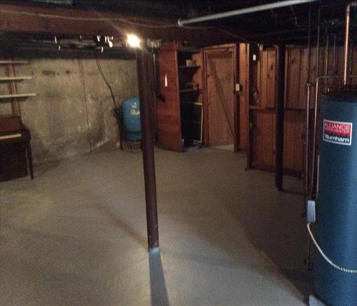 Unfinished basement piano in left corner, blue water heater. Half the walls are wood panels & other half concrete with shelf